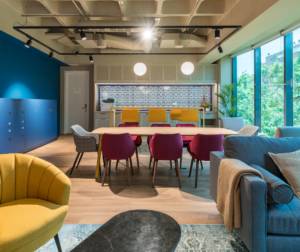 Sala de colaboración en oficina presencial, con mobiliario de colores amarillo, azul y rosa, iluminación natural por medio de ventanas y equipamiento para zona de reuniones.
