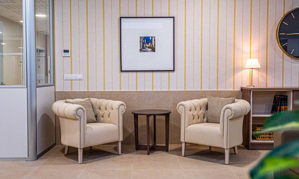 Sala de espera con mobiliario blanco dos butacas, mesa y almacenamiento con pared con papel pintado