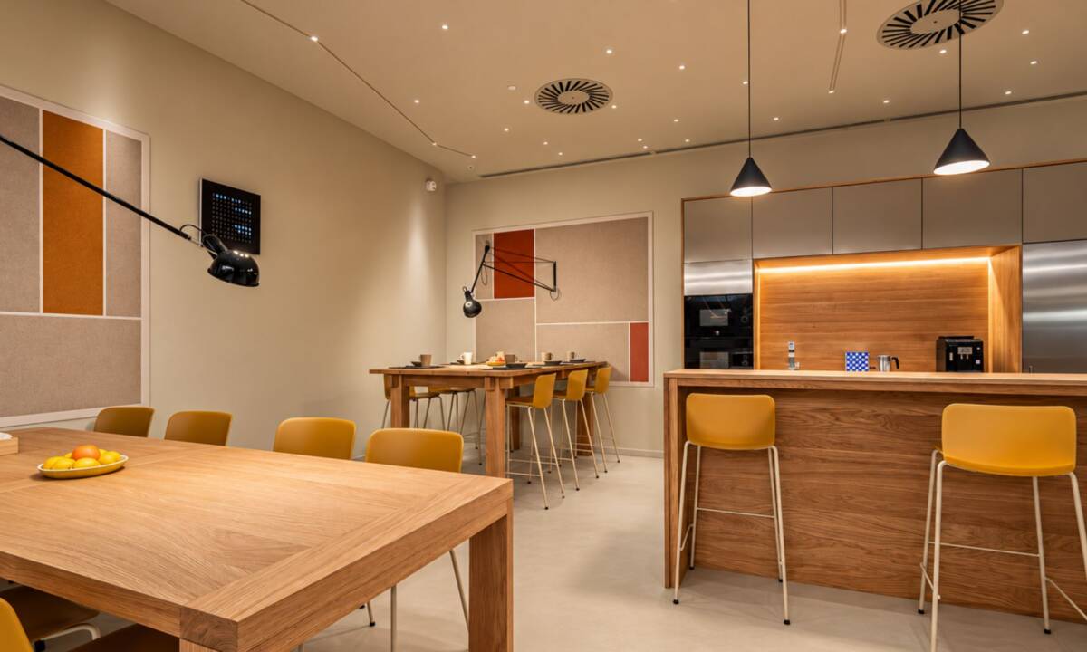Espacio de cantina con mobiliario a medida de roble, sillas en color mostaza, mobiliario de cocina en inox y paneles acústicos