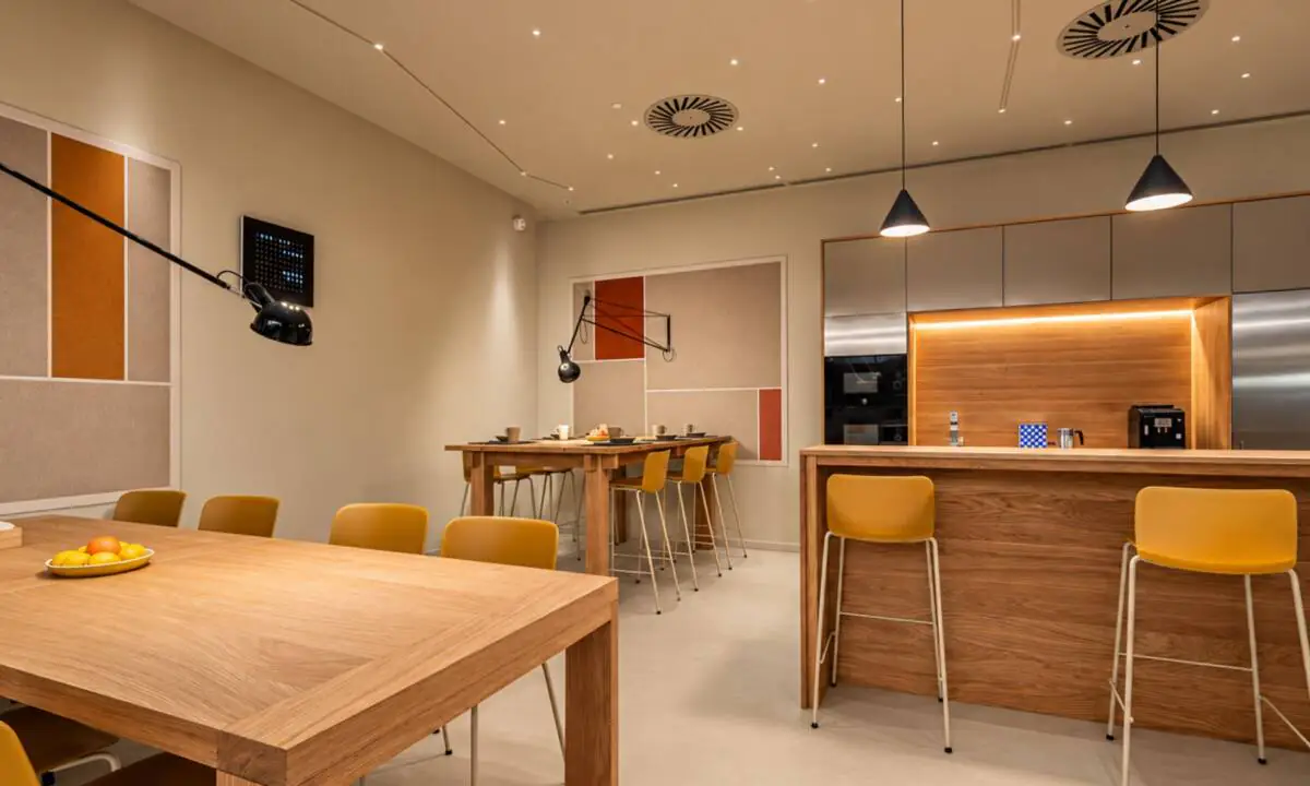 Espacio de cantina con mobiliario a medida de roble, sillas en color mostaza, mobiliario de cocina en inox y paneles acústicos